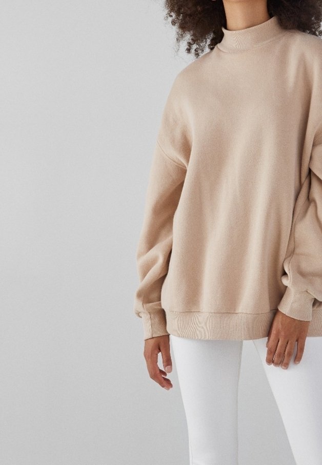 Beige tröja med fuskpolokrage, långa voluminösa ärmar och ribbad fåll. Mjuk tygstruktur; bärs med vita figursydda byxor.