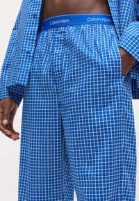 Set de pijama în carouri albastre, cu o textură moale. Bluza are un design cu nasturi, în timp ce pantalonii dispun de un elastic cu marca Calvin Klein.
