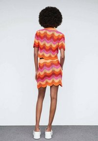 Femme avec une coiffure afro portant un haut en crochet aux motifs ondulés colorés orange, rose et rouge ainsi qu'une jupe assortie, se tenant devant un mur blanc.