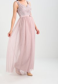 Lange, vloeiende jurk met een sheer, lichtroze rok en een kanten lijfje met bloemmotieven. Heeft een aansluitende taille en schouderbanden.