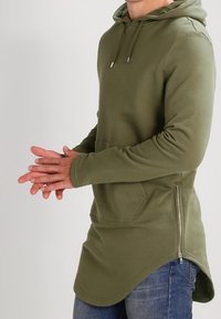 Sudadera con capucha de color verde olivo, de manga larga, con bolsillo tipo canguro y detalles de cremallera lateral. La tela tiene una apariencia suave, con un corte relajado y un dobladillo curvado.
