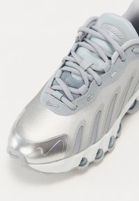 Sapatilha atlética prateada com parte superior em malha, biqueira arredondada e sola almofadada com unidades de ar visíveis, atacadores cinzas detalhados e acentos de logotipo.