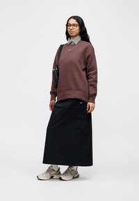 Μπορντό φούτερ Nike με στρογγυλή λαιμόκοψη, μαύρη φούστα cargo και μπεζ αθλητικά παπούτσια. Γκρι πουκάμισο με γιακά φαίνεται κάτω από το φούτερ.