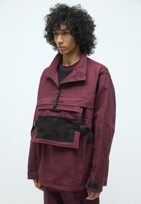 Burgunderfarget genserjakke med høy krage, frontlomme i mesh, justerbare svarte stropper og avslappet passform. Materiale laget av en blanding av bomull og polyester.