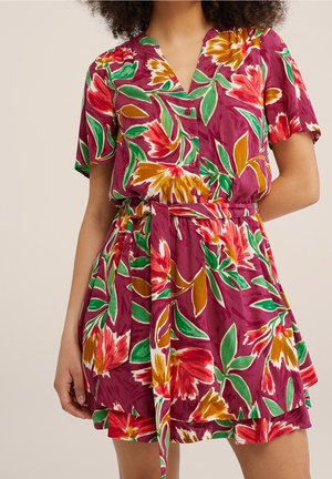 Kvinde iført en kortærmet kjole med knapper og et blomsterprint i rød, grøn, gul og hvid på en dyb magenta baggrund.