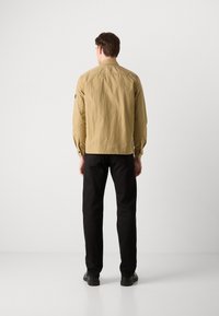 Veste légère beige avec une finition texturée, des poignets boutonnés et un logo sur la manche, associée à un pantalon noir à jambes droites et des chaussures noires.