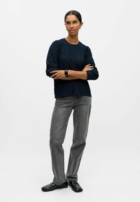 Pull en maille texturée bleu marine avec des manches bouffantes, associé à un jean droit gris et des mocassins noirs. Le modèle pose les bras croisés.