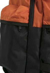 Ryggsekk med en oransje toppdel med quiltet tekstur, supplert med en svart nederdel, har to glidelåser og sidestraps.