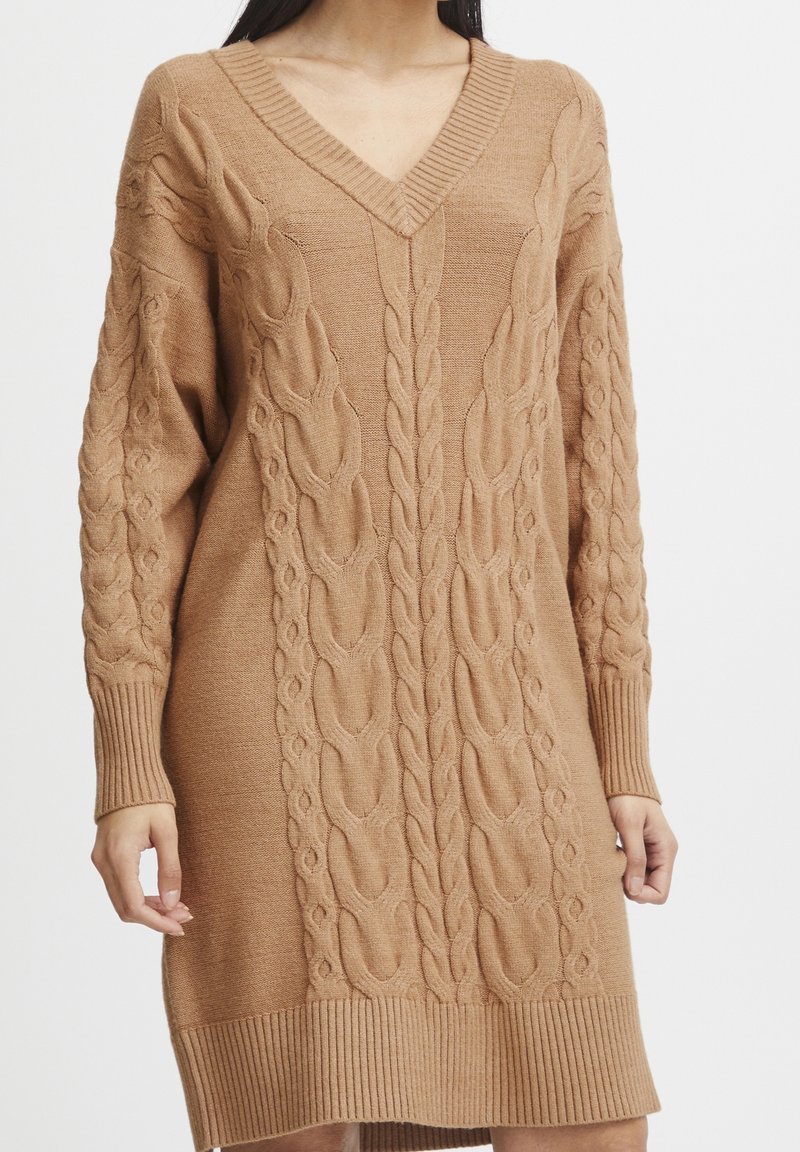 Femme portant une robe-pull camel à manches longues, en maille torsadée, col en V avec poignets et ourlet côtelés.