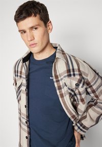 Plaid shirt in beige, brown, and navy over a navy T-shirt with pocket. Features button-up design and stitched accents. Smooth texture.