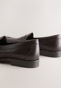 Paire de chaussures habillées en cuir marron foncé à enfiler avec de petits talons en caoutchouc noir sur un fond clair uni.