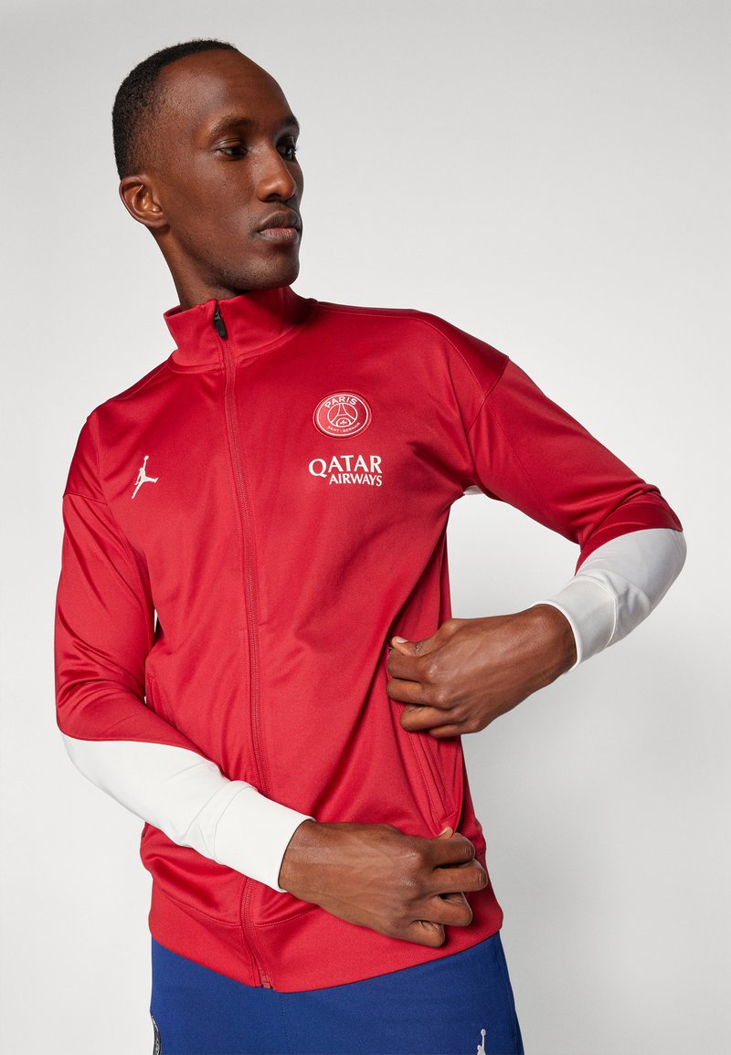 Chaqueta deportiva roja con cuello alto y cierre de cremallera en la parte frontal. Acentos blancos en las mangas, que presentan el logo de París y la marca de Qatar Airways. Tejido suave.