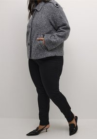 Grå tekstureret jakke med krave, to forlommer og sorte knapper, kombineret med sorte tætsiddende bukser og sorte spidse hæle.