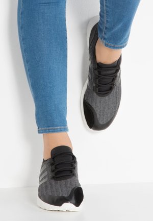 Nohy v modrých džínách s černo-šedými teniskami s bílými podrážkami, stojící před bílou zdí a podlahou.