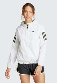 Bela lahko jakna s kapuco, ki ima črne tričrte na rokavih in sprednji zadrgo.