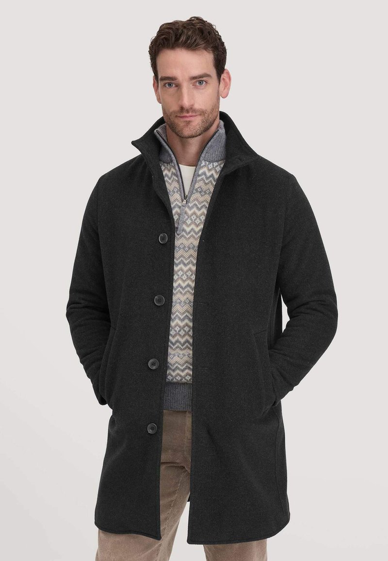 Dunkelgrauer, knöpfbarer Mantel mit hohem Kragen aus strukturiertem Stoff. Wird über einem gemusterten Pullover getragen und verfügt über seitliche Taschen.