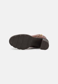 Botte en cuir marron avec une surface texturée, bout rond et une semelle épaisse en caoutchouc noir. Accent de zipper doré sur le côté.