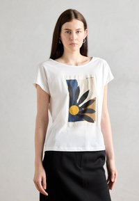 T-shirt blanc à manches courtes, arborant un imprimé graphique de feuilles bleues et noires, avec un cercle jaune, sur un fond beige.
