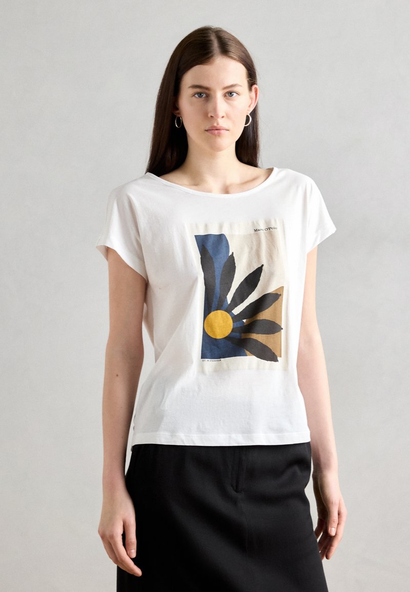 T-shirt blanc à manches courtes, arborant un imprimé graphique de feuilles bleues et noires, avec un cercle jaune, sur un fond beige.