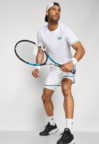 Valge tennisekomplekt, millel on lühikeste varrukatega särk roheliste aktsentidega ja lühikesed püksid. Mudel hoiab patsi, kandes musti jalatseid ja randmeääriseid.