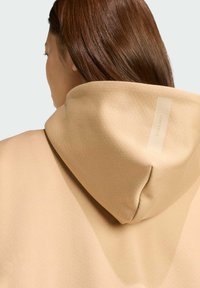 Beige hoodie van een gestructureerde stof, met een grote capuchon en een label op de achterkant met de tekst "SPORTSWEAR."