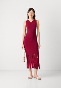 Dopasowana bordowa sukienka dzianinowa z okrągłym dekoltem, z otwartymi rozcięciami na końcu i frędzlami. W połączeniu z jasnym torebką i sandałami.