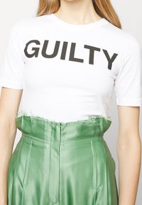 Camiseta de algodón blanca con la palabra "CULPABLE" en letras negras en negrita, combinada con pantalones de satén verdes de talle alto con un dobladillo desflecado.