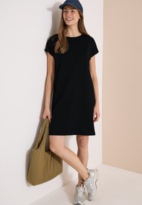 Femme portant une robe noire à manches courtes et une casquette bleue, tenant un grand sac vert olive, debout avec une jambe croisée, souriante.