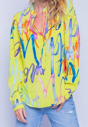 Donna che indossa una camicetta a maniche lunghe di un giallo brillante con motivi astratti colorati e jeans azzurri strappati.