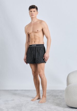 Man zonder shirt, blootsvoets staand op een grijs tapijt, wearing zwarte boxer shorts met patroon van Lacoste tegen een lichtgrijze achtergrond.