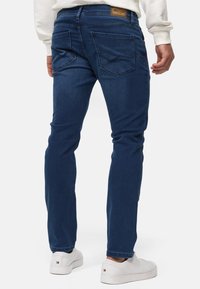 Mørkeblå denimjeans med slim fit. Har to baklommer med sømdetaljer og en etikett på linningen. Brukt med hvite joggesko.