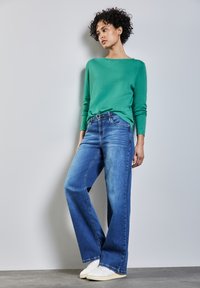 Grüner Strickpullover mit langen Ärmeln, kombiniert mit locker sitzenden blauen Jeans. Die Jeans haben ein klassisches Fünf-Taschen-Design und ein gerades Bein.