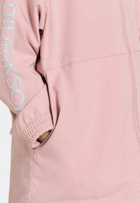 Rosa jacka med dragkedja, elastiska ärmmuddar och sidofickor. Varumärkeslogotyp tryckt i silver längs ärmen. Slät tygtextur.