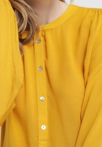 Blouse jaune avec un col rond, dotée d'une fermeture à boutons à l'avant avec des boutons en argent et de longues manches bouffantes. Tissu doux et texturé.