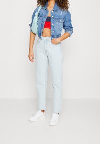 Mujer que lleva jeans de tiro alto en un tono azul claro, chaqueta de mezclilla azul corta, top tubular rojo y azul marino, zapatillas blancas y un bolso de hombro en azul claro.