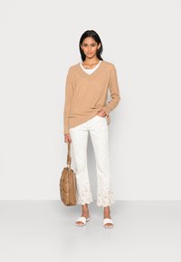 Suéter bege com decote em V, blusa branca em camadas por baixo, calças brancas com padrão e bordado de renda, segurando uma bolsa bege, usando sandálias brancas.