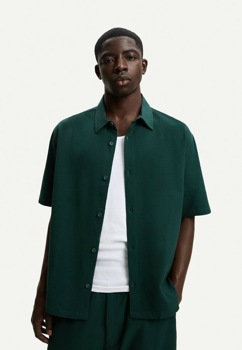 Short-sleeve button-up shirt in dark green, featuring a textured fabric, pointed collar, and visible buttons, worn over a white tank top.