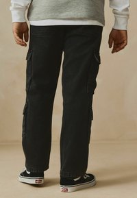 Pantalons cargo noirs avec poches latérales, coupe décontractée et une texture légèrement délavée, associés à des baskets Vans noir et blanc.