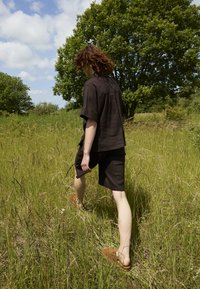 Personne aux cheveux ondulés portant une chemise noire et un short, marchant pieds nus dans une herbe haute dans une prairie ensoleillée avec des arbres verts.