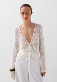 Pulover alb din tricot, cu decolteu în V adânc, sistem de șnur la talie, mâneci lungi și un model complicat de dantelă pe întreaga suprafață.