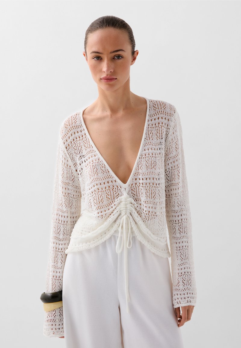 Pulover alb din tricot, cu decolteu în V adânc, sistem de șnur la talie, mâneci lungi și un model complicat de dantelă pe întreaga suprafață.