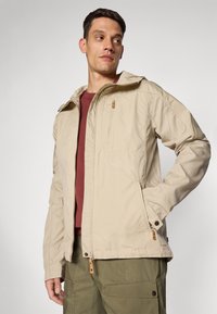 Veste extérieure beige avec capuche, poche zippée et accents en bouton. Portée sur une chemise bordeaux, associée à un pantalon cargo vert olive.