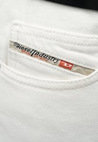 Ύφασμα λευκού τζιν με μια μικρή τσέπη που φέρει μια μπεζ ετικέτα με το κείμενο "diesel industry denim division" σε μαύρο και κόκκινο χρώμα.