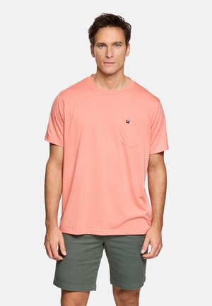 Hombre con camiseta de manga corta color coral con bolsillo en el pecho y pantalones cortos de longitud hasta la rodilla en verde oliva, de pie contra un fondo liso.