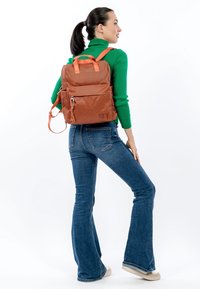 Bruine rugzak met textuur, met oranje riemen en accenten, voorzien van een voorvak en logo-detail, gedragen door een model met wijde jeans.