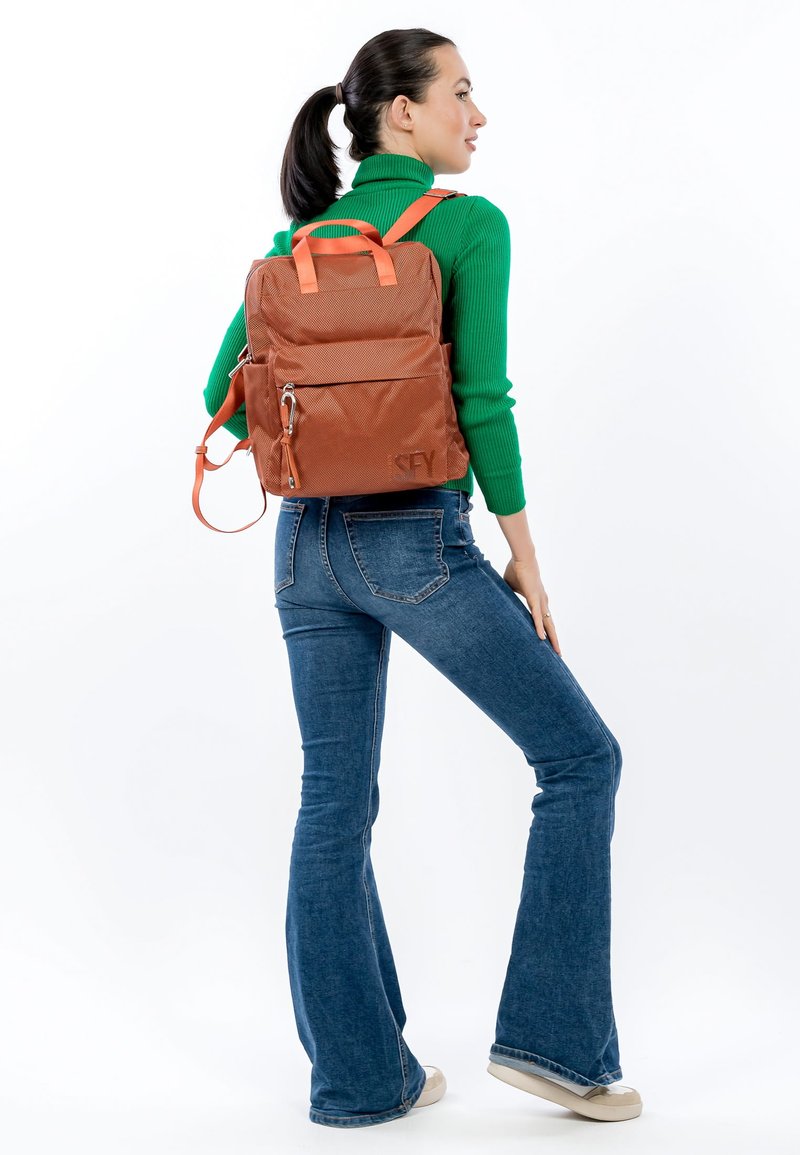 Bruine rugzak met textuur, met oranje riemen en accenten, voorzien van een voorvak en logo-detail, gedragen door een model met wijde jeans.