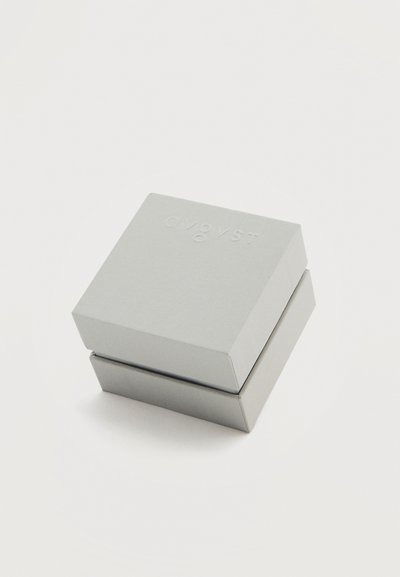 Μικρό, κλειστό τετράγωνο κουτί κοσμημάτων με διακριτικό ανάγλυφο όνομα μάρκας στην κορυφή, τοποθετημένο σε απλή λευκή επιφάνεια.