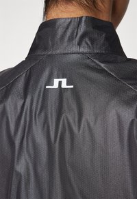 Svart tygjacka med hög krage och synlig ripstop-textur, med en vit logotyp centrerad under kragen på baksidan.