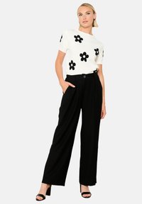 Top in maglia bianca con applicazioni di fiori neri, abbinato a pantaloni neri larghi. La modella si posa con le mani nelle tasche dei pantaloni, indossando tacchi neri.