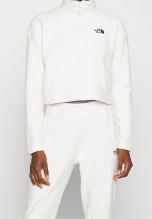 Personne portant un sweat-shirt court blanc zippé et un pantalon de survêtement blanc assorti avec les logos The North Face sur les deux vêtements.